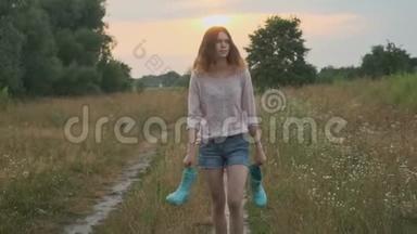 小女孩走在农村的路上，手里拿着靴子。 穿脏衣服的快乐孩子
